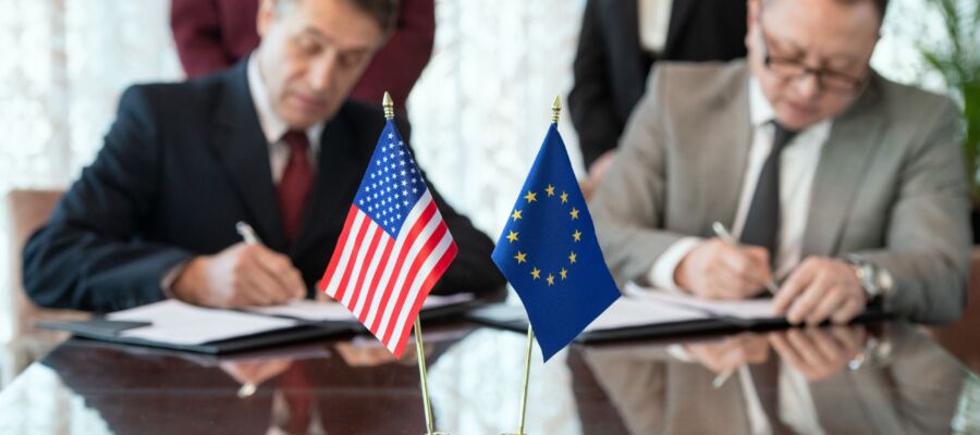 Flags of United States and European Union on table against two delegates signing contract after negotiating and making deal