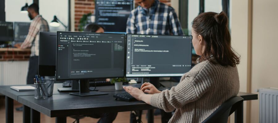 Focused software developer writing code looking at multiple computer screens displaying machine learning algorithm. Programer coding user interface while colleagues doing teamwork in background.